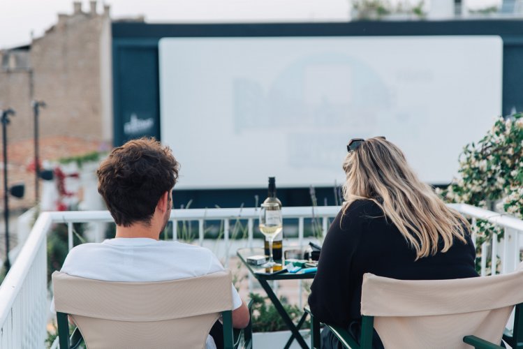 a man and a woman sitting on white chairs looking at a screen at an open-air cinema