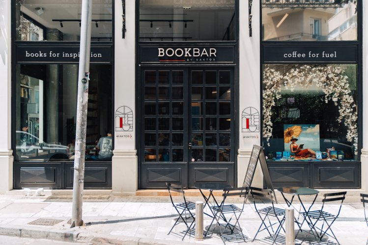 A street-level view of a bookstore café with black-framed windows, signs for books and coffee, and a couple of outdoor tables and chairs.