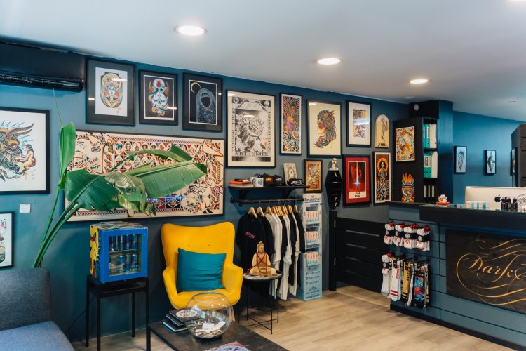 A tattoo studio with a blue wall adorned with framed tattoo art. There is a yellow armchair, a small table, and a display of merchandise.