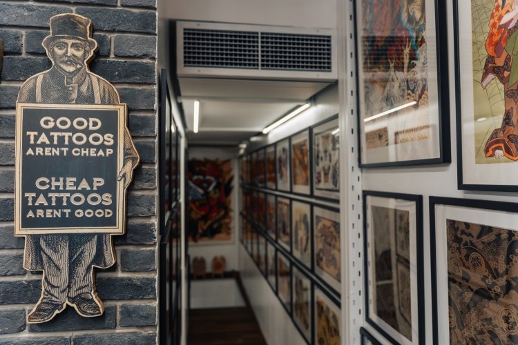 A narrow hallway in a tattoo shop with framed tattoo designs, a sign on the wall reads "Good Tattoos Aren't Cheap. Cheap Tattoos Aren't Good."