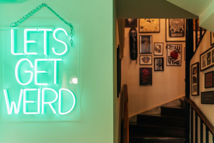 A neon sign that reads "Let's Get Weird" hanging on a wall. Behind it, a staircase leads up to a room filled with framed artwork.