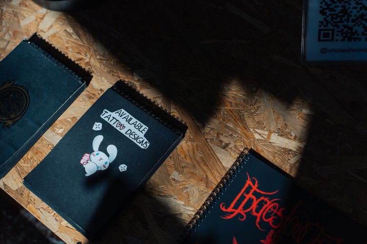 A photo of three black sketchbooks on a wooden surface. One of the sketchbooks has the text "AVAILABLE TATTOO DESIGNS" written on it.