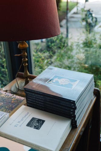 A stack of books titled "Wings of Thought" rests beside a classic brass lamp with a red lampshade, placed near a window with greenery visible outside.