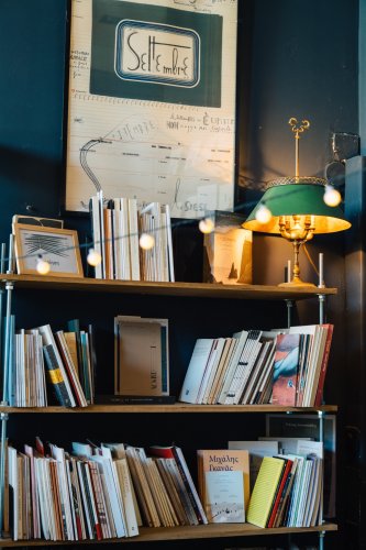 A cozy bookshelf filled with a variety of books, illuminated by warm string lights and a vintage green lampshade, set against a dark blue wall.