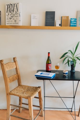 A rustic wooden chair placed beside a small black table with a bottle of wine, a glass, and a bowl of nuts, with a plant and books displayed on a wall shelf in the background.