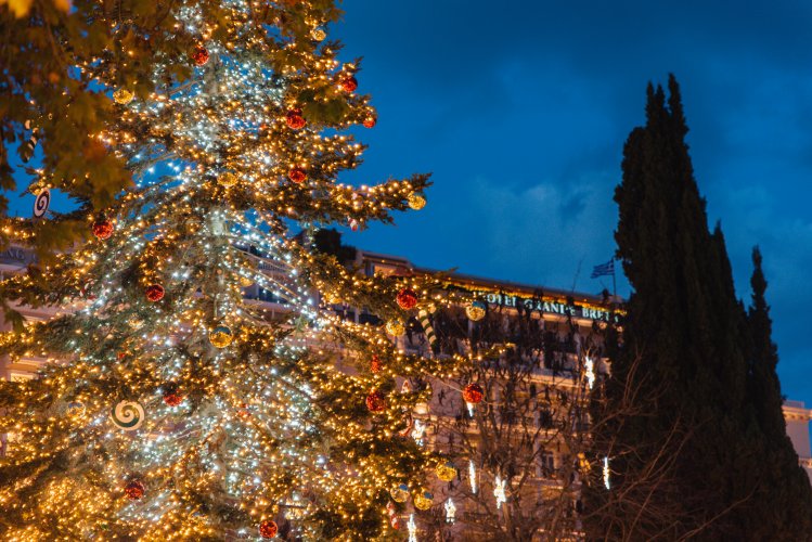 A tall Christmas tree adorned with colorful lights and ornaments stands in a square. The Grand Bretagne hotel is visible in the background.