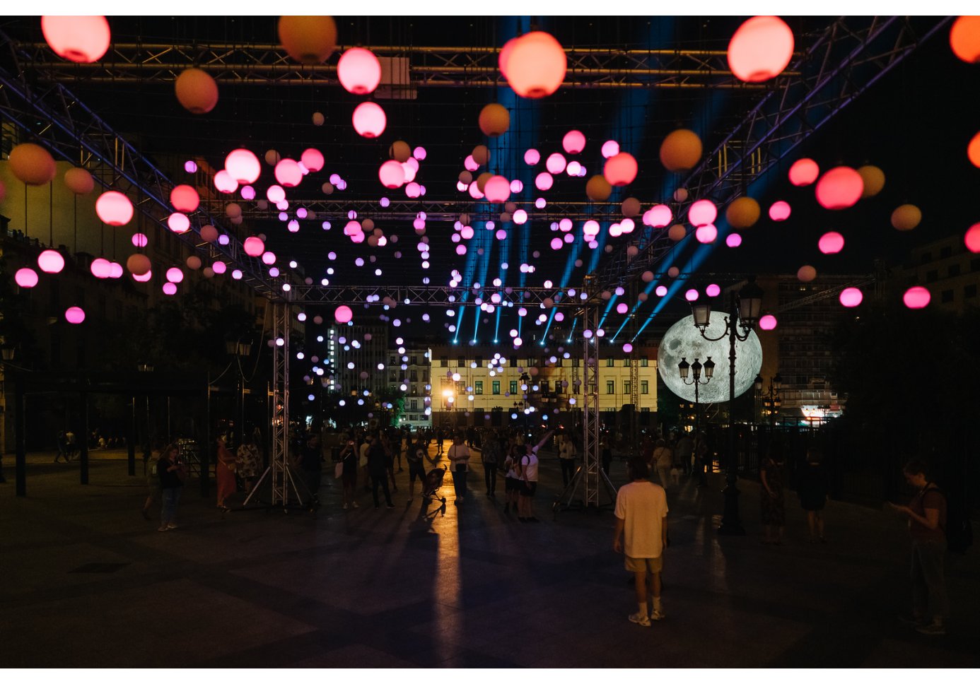 people walking under an art installation with many lights and a moon in a square, nighttime.