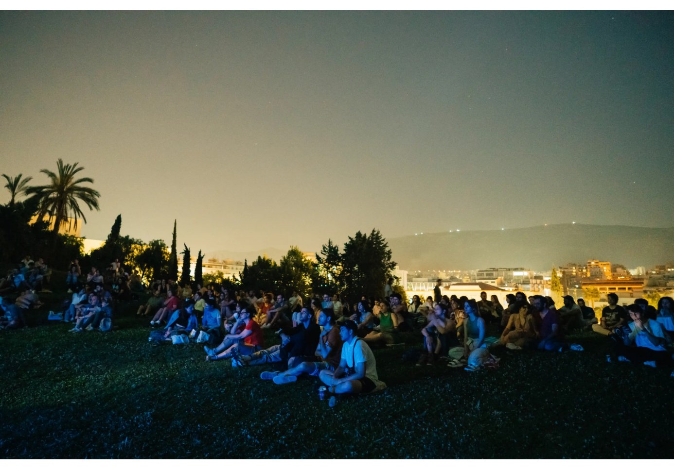 people on a hill watching a movie, city lights behind them