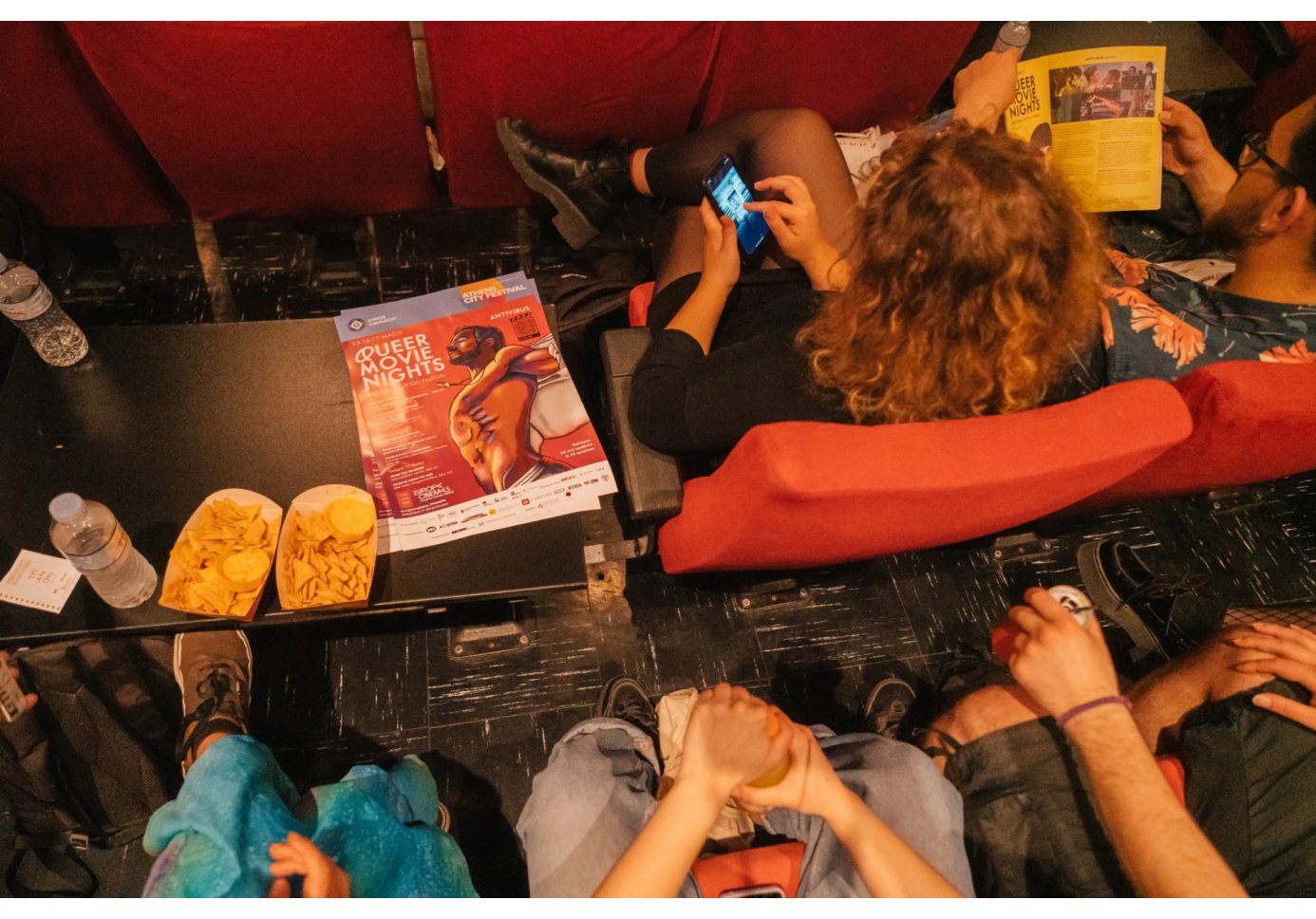 people sitting in a movie theatre, nachos and a programme that reads "queer movie nights" around