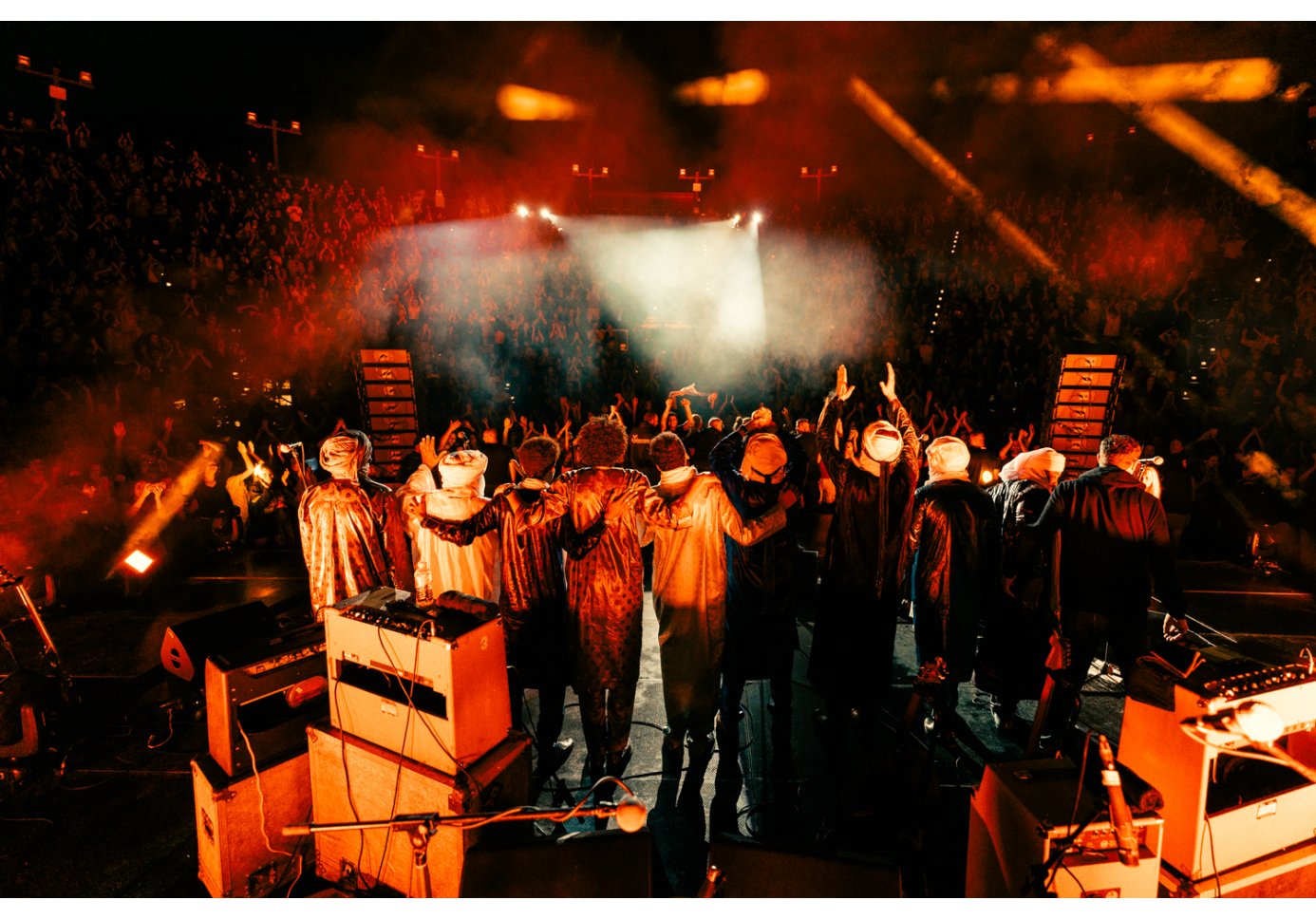 a band standing on stage with their backs to the camera, facing an enthusiastic audience in a large open-air concert venue