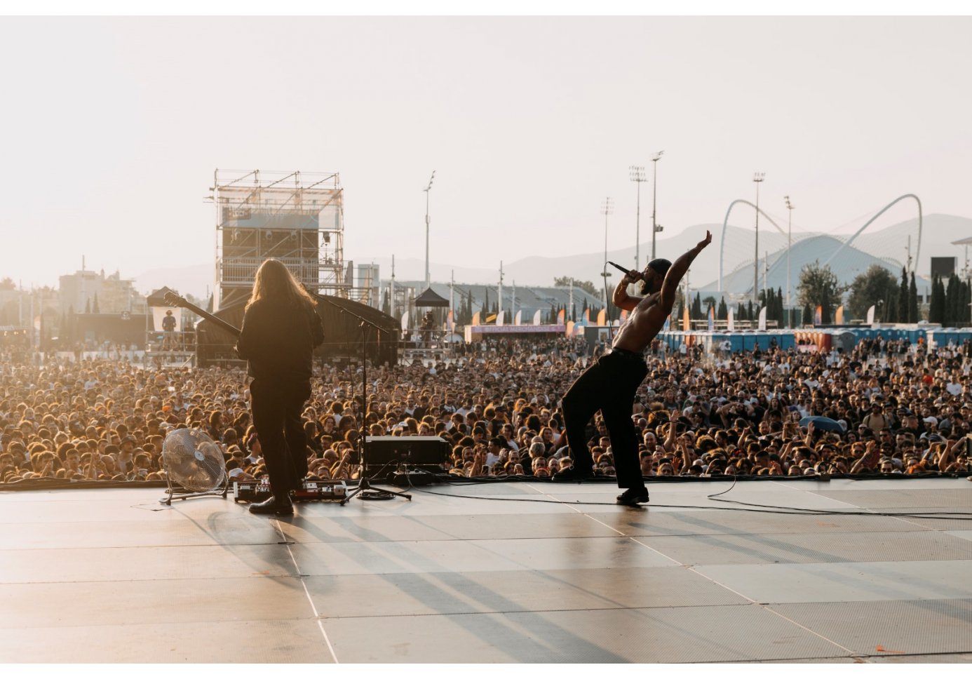 A live outdoor concert captured from the stage, showing a shirtless vocalist performing, beside him, a guitarist with long hair 